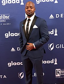 A handsome Black man in a blue suit standing in front of an awards ceremony backdrop.