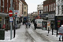 Street scene with pedestrians and vehicles in road lined with shops. There is snow on the ground.