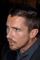 Profile shot of a man looking down and to his right. He has a goatee and stubble, and is wearing a black dress shirt with collar and a black blazer.