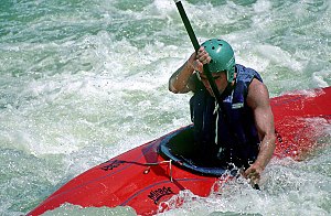 Man wearing helmet sitting in a fiberglass boat, paddling through frothy water