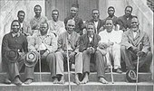 A black and white photo of 13 Christian Zulu men seated outside of a church.