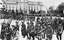 Schwarzweißfotografie von einer marschierenden Armee mit Gewehren auf den Schultern. Im Hintergrund sind eine Menschenmenge und ein großes Gebäude.