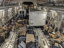 Photograph of the inside of a cargo jet, containing people in military field uniforms, equipment, and trucks