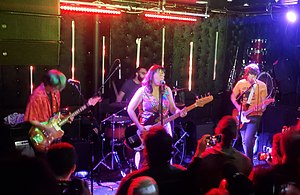 Onstage at a small crowded venue, left to right: Katie Park playing guitar, Daoud Tyler-Ameen in the back playing drums, Emma Cleveland playing bass, and David Combs playing guitar