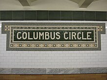 A mosaic plaque on the IRT platform with the words "Columbus Circle" in capital letters, surrounded by orange-tinted tiles. There is a white tiled wall below the plaque.