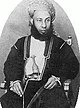 A black-and-white photograph of a man with a dark beard wearing a turban, a dark jacket, and a white shirt, sitting, and looking at the viewer