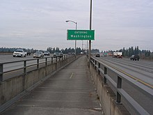 View of a narrow concrete pathway between freeway lanes, separated by a concrete barrier topped with metal railings.