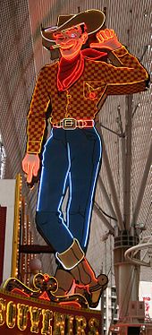 Photograph of a large painted sign in the form of a cowboy. The cowboy is winking his eye. His left hand is lifted, and he's pointing that thumb towards the building to his right. A lighted cigarette dangles from the corner of his mouth. He's wearing a cowboy hat, boots, and a scarf. Glowing neon tubes highlight the outlines.