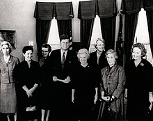 Seven women and President Kennedy standing in the Oval Office in 1962.