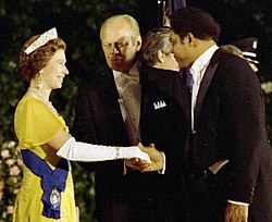 Photograph of Queen Elizabeth II wearing a gold gown with a purple sash across it and a crown on her head smiles at Mays dressed in a tuxedo, while in the center of the photo, Gerald Ford wearing a tuxedo with white bowtie looks at the Queen.