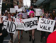 A group of people walk in a demonstration (a SlutWalk) in New York City; many of them carry signs with slogans relating to and condemning sexual assault and discussing public perceptions thereof