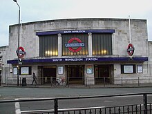 A wide two-storey stone-faced building has three square entrances at the centre beneath a dark blue awning with the words "SOUTH WIMBLEDON STATION". Above the awning is a wide glazed screen in three panels, the centre one of which contains the Underground roundel of a red ring with a blue bar and the word "UNDERGROUND". Two smaller roundels either side of the entrances are mounted on poles at right angles to the face of the building.