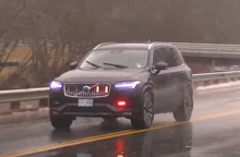 A new, black Volvo SUV driving in the rain, with red emergency lights activated while a firefighter responds to a call