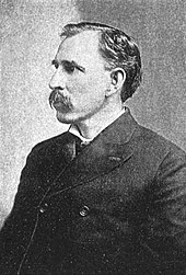 A man in his late forties with black hair and a mustache. He is wearing a tightly buttoned black coat and facing left.