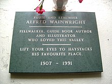 black stone plaque reading: Pause and remember Alfred Wainwright: Fellwalker, guide book author and illustrator who loved this valley. Lift you eyes to Haystacks his favourite place. 1907-1991