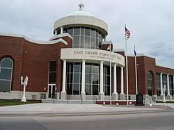 A red brick building with a glass lobby. A memorial sign and flags are located in front of the building.