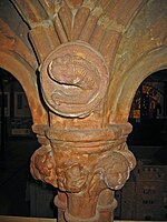The top of a column and base of two arches. In the spandrel is a carving of an otter-like creature; the capital is decorated with a carved head and foliage