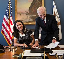 Julia Louis-Dreyfus seated at a desk, with Biden standing over her, both laughing.