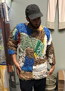 A man in a hat wears a colorful sweater knit from plastic yarn. It is patchwork. he appears to be standing in a clothing store.