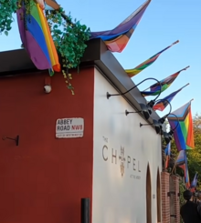 A building has a red wall with a sign that reads "Abbey Road NW8". The other wall is tan, and painted on it is "The Chapel at the Abbey". Progress pride flags are hung from the top of the building.