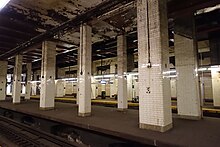 View of the Chambers Street station's central island platform from the easternmost island platform. The station has a high ceiling, and there are tiled beams supporting the Municipal Building above.