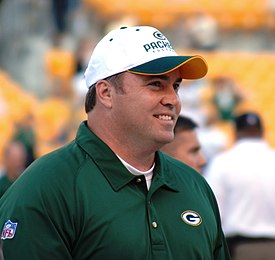 Candid photograph of McCarthy wearing a green Packers polo shirt and white Packers baseball cap in a football stadium
