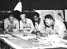 Black and white photograph of four men wearing military uniforms working on maps which have been placed on a table