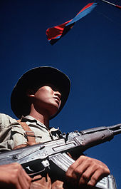 Looking from the waist up, a man wearing a hat and holding an assault rifle with one hand holding the magazine and the other on the pistol grip