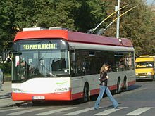 Frontale Farbfotografie eines rot-weißen Busses mit der Anzeige „16 Pasilaiciai“, der vor einem Zebrastreifen hält und Fahrgäste einsteigen lässt. Der Bus ist durch zwei Stangen mit der Oberleitung verbunden. Rechts läuft eine Frau über den Zebrastreifen. Im rechten Hintergrund steht ein gelber Kleintransporter.