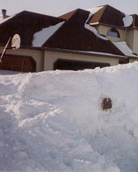 A large pile of snow piled up in front of a home, nearly obscuring it. A mailbox is peeking through the pile of snow and a basketball hoop can be seen behind the snow in front of the home. Snow is piled up in small areas on top of the home's roof.