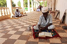 A dark-skinned boy sits cross-legged on a small mat on a tiled floor in a shaded collonade. He is wearing light clothes and a white cap and is looking down at a book written in Arabic. Beside him is a tin can. Some distance behind him are more boys sitting and standing.