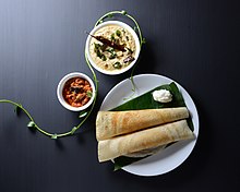 Two dosa rest next to a dollop of butter on a plantain leaf. There are separate bowls for the sauces.