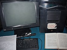 Fotografie. Zeigt einen Monitor, einen Rechner und eine Tastatur. Die Geräte sind gekennzeichnet mit „PROPRIETE CERN“ und „NeXT“. Auf dem Rechner klebt ein teilweise durch Beschädigungen unleserlicher Sticker mit dem handschriftlichen Text „This machine is a ser [...] DO NOT POWE [...] DOWN!!“ Auf der Tastatur liegt ein Ausdruck von „Information Management: A Proposal“, daneben ein aufgeschlagenes Buch.