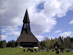 Foto einer dunkelbraunen Holzkirche mit einem Holzturm. Um die Kirche herum befindet sich ein Friedhof