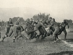 A cluster of American football players collide on a dirt field