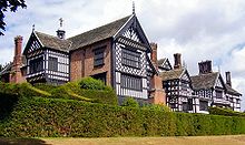 The exterior of a large house. There are several chimneys, leaded windows and wings. In the foreground are two rows of hedges.