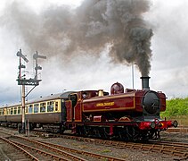 A pannier tank locomotive is pulling two cream and brown passenger carriages. The locomotive is mainly painted maroon, but there is black and yellow lining on the side of the pannier tank, the cab, the bunker, the toolbox, and the splashers. The words "LONDON TRANSPORT" are shown in yellow on the side of the pannier tank. The chimney and front of the smokebox are black. The front buffer beam is red.