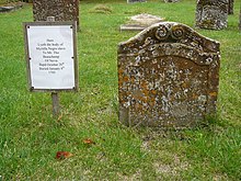 A small stone headstone, dotted with lichen, in a graveyard. The headstone has a simple carved frame, with a scroll design at the top, and an inscription in the Roman alphabet. Next to the headstone is a sign with a transcription of the inscription, which reads "Here lyeth the body of Myrtilla, negro slave to Mr. Thos Beauchamp of Nevis. Bapt. Oct. ye 20th. Buried Jan ye 6th, 1705"