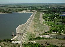 From the west, a large dam and the body of water it holds sit to the left of a grassy area with some trees