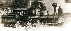 A steam locomotive with a tender parked on a railroad track. Three railroad workers pose in front of it, along with a fourth in the locomotive's cab.