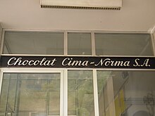 Dieses Bild zeigt einen kursiv gestalteten Schriftzug der Chocolat Cima-Norma S.A. (weiss auf dunklem Grund), der über einer Glastür angebracht ist.