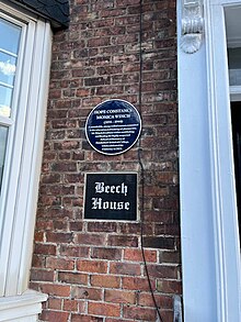 A commemorative round blue plaque on a brick building with white wooden windows and columns. An additional sign reads Beech House.