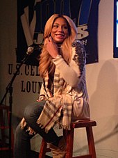 An image of a blonde woman sitting on a stool in front of a microphone. She is looking away from the microphone.