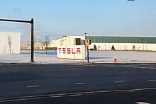 Large white sign, with red letters spelling "Tesla", in the parking lot of a long, low building.