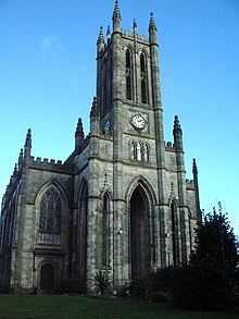 A large church seen from the northwest, showing a tower with clock faces over arches, an arched window, and crocketted pinnacles.