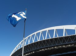 A blue flag with a white number 12 flies against a clear sky. An expansive white roof truss is behind the flagpole.