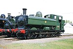 A green pannier tank locomotive, viewed from the left and front, is standing in front of two other locomotives. The locomotive is fitted with a chimney, rounded boiler dome, and a curved safety valve cover. The cab is enclosed with rectangular windows. Although mainly green, the chimney, front, and undercarriage are painted black, except the coupling rods which are grey, and the front buffer beam is painted red. On the side of the pannier tank is a yellow roundel with the letters G W R. On the side of the cab there is a small blue disc and a black number plate with the letters and edging in yellow.