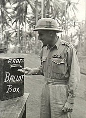 Three-quarter-length outdoor portrait of moustacioed man in light tropical military uniform with pith helmet and pilot's wings on left breast pocket, placing folded paper into a box marked "RAAF BALLOT BOX"