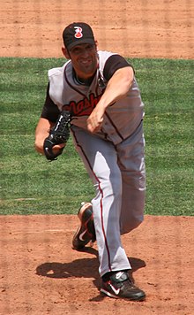 A man in a gray baseball uniform with "Nashville" written on the chest and a black cap with a red and white music note on the front leaning forward on the pitcher's mound with his left arm out has just thrown a ball.