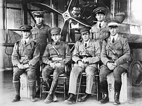 Six men in military uniforms with peaked caps, two standing and four seated, in front of a biplane